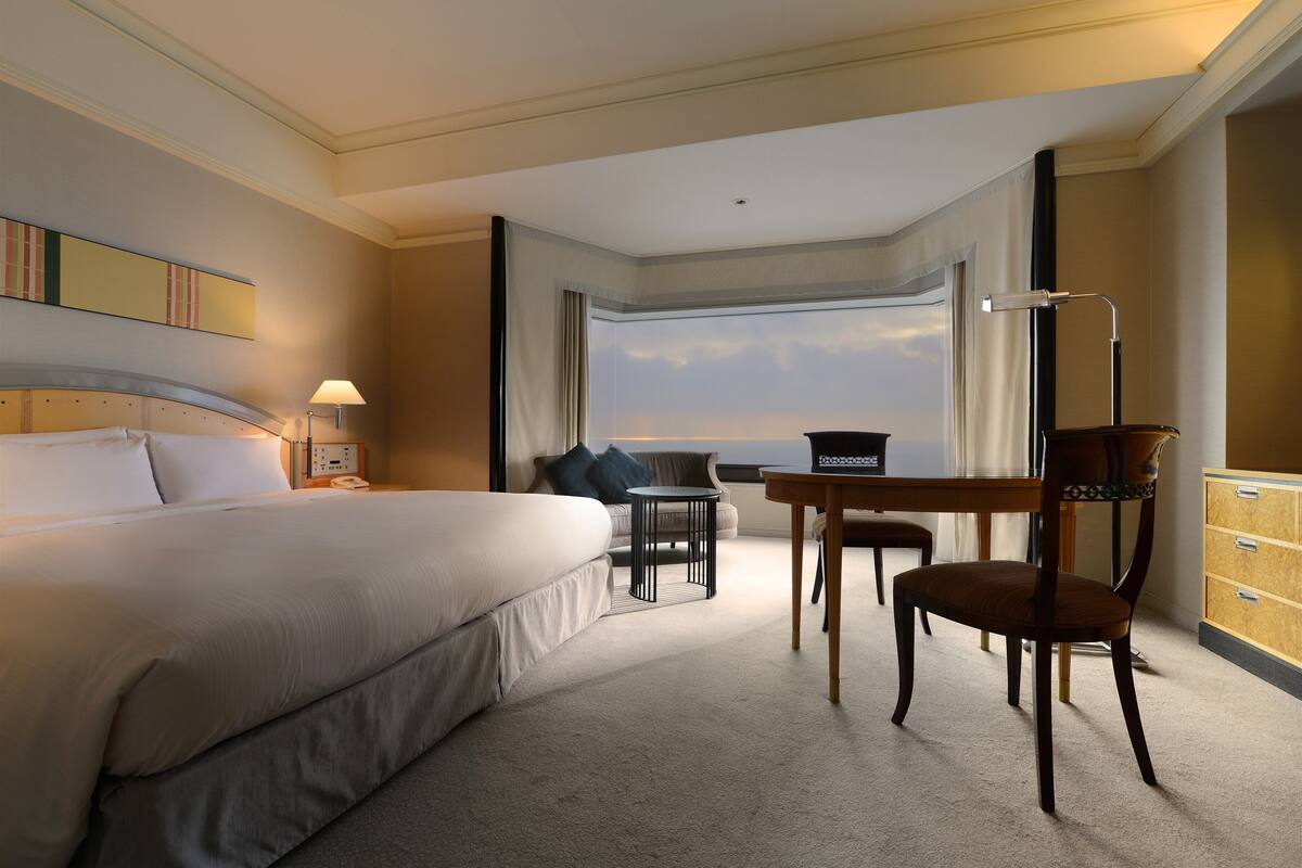 Een hotelkamer met een groot bed, een bureau met stoel, een lamp en uitzicht op de lucht door een raam.