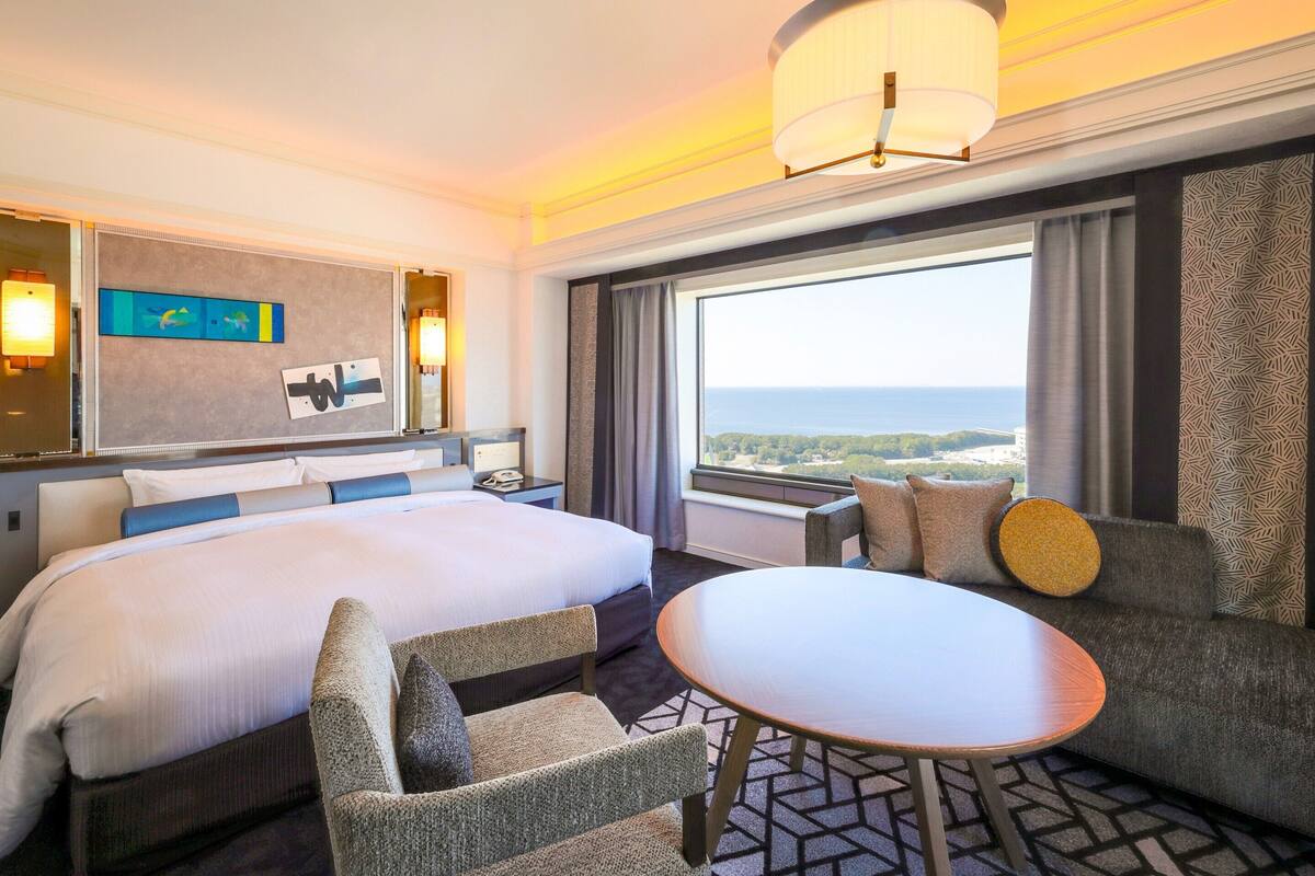 Hotelkamer met een bed, een bank, een kleine tafel, een groot raam met uitzicht en een kroonluchter.
