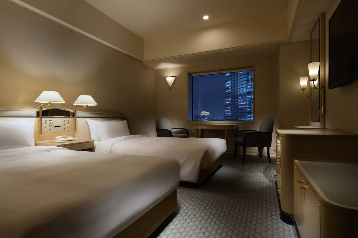 Een hotelkamer met twee bedden, een bureau, een stoel en een groot raam met uitzicht op de stad.