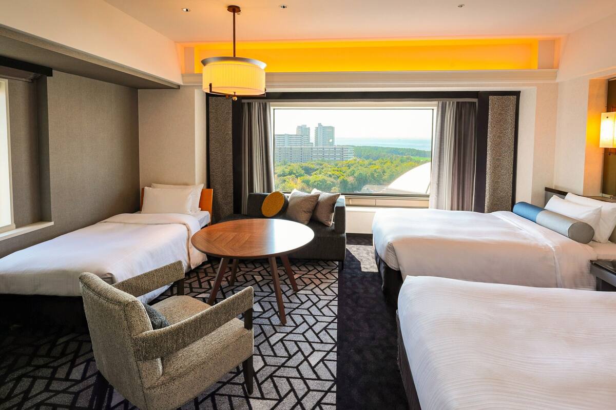 Een hotelkamer met twee bedden, een bank, een ronde tafel en uitzicht op een stadsbeeld.