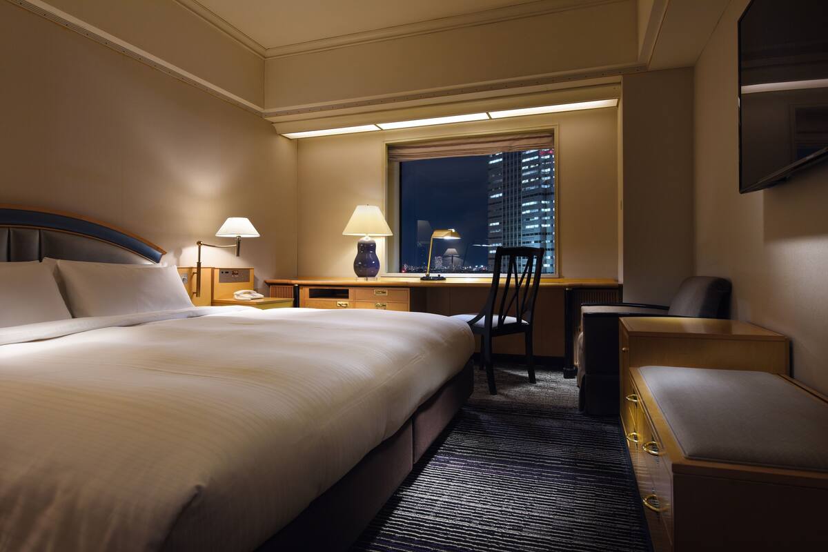 Hotelkamer met een groot bed, een bureau met lamp, een stoel en uitzicht op een stadsgezicht door een raam.