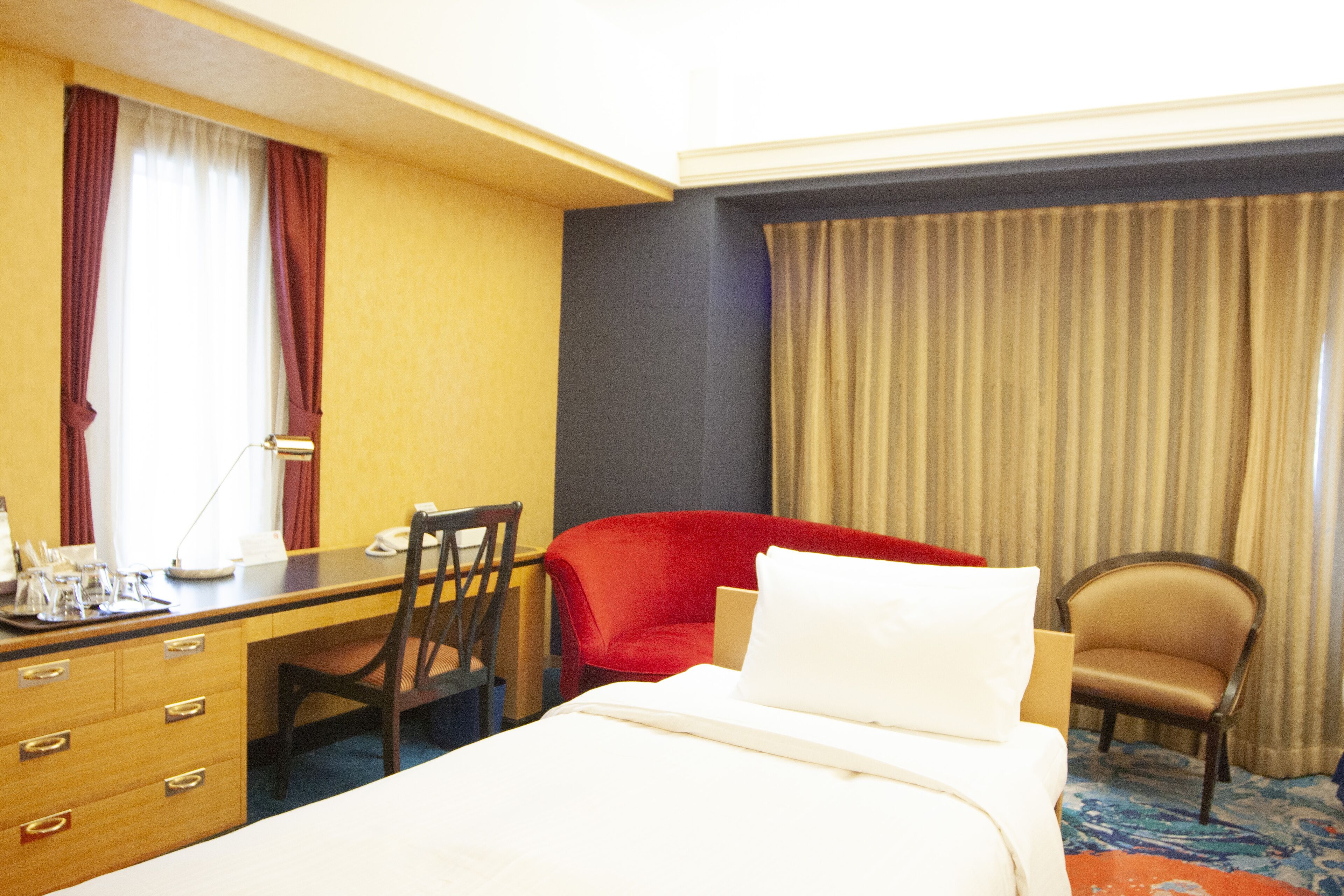 Una habitación de hotel con cama, escritorio, una silla roja, una silla marrón y ventana con cortinas.