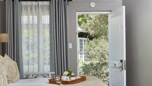 A bedroom with a large bed, a chandelier, a window with curtains, and a door leading to a garden. - The Devon Valley Hotel (Stellenbosch)