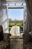 Puerta abierta que da a un patio con una mesa para dos, una silla y vistas a follaje.
