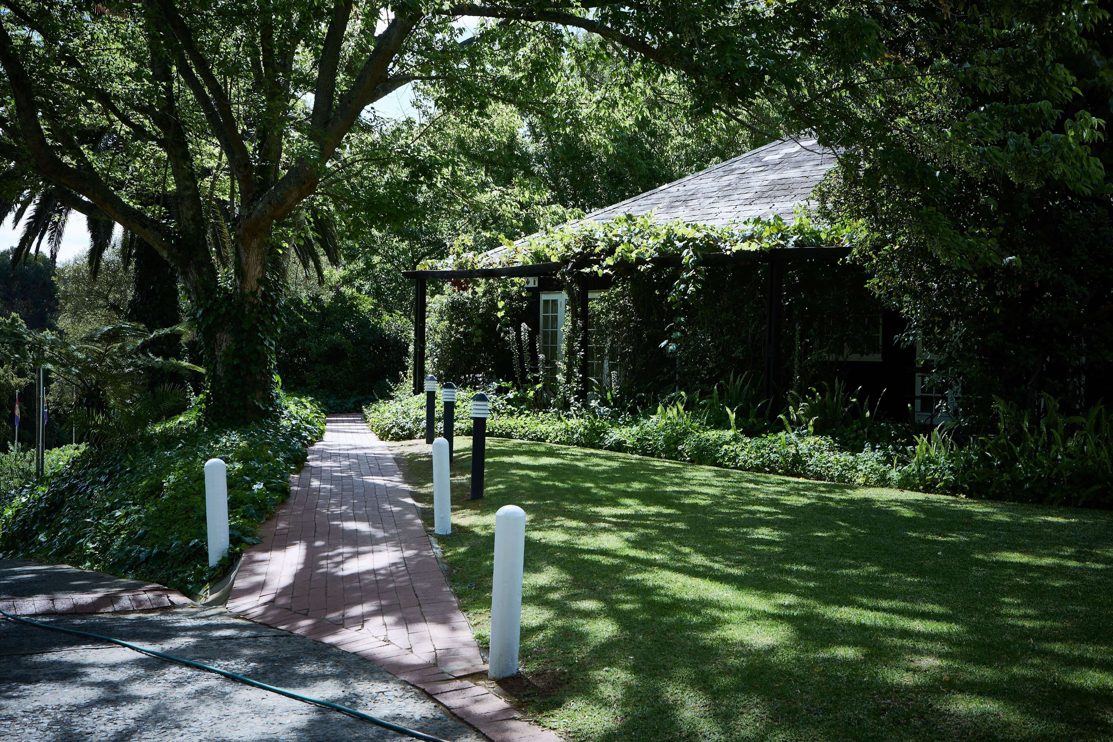 A garden pathway leading to a gazebo with a tiled roof, surrounded by lush greenery and trees.