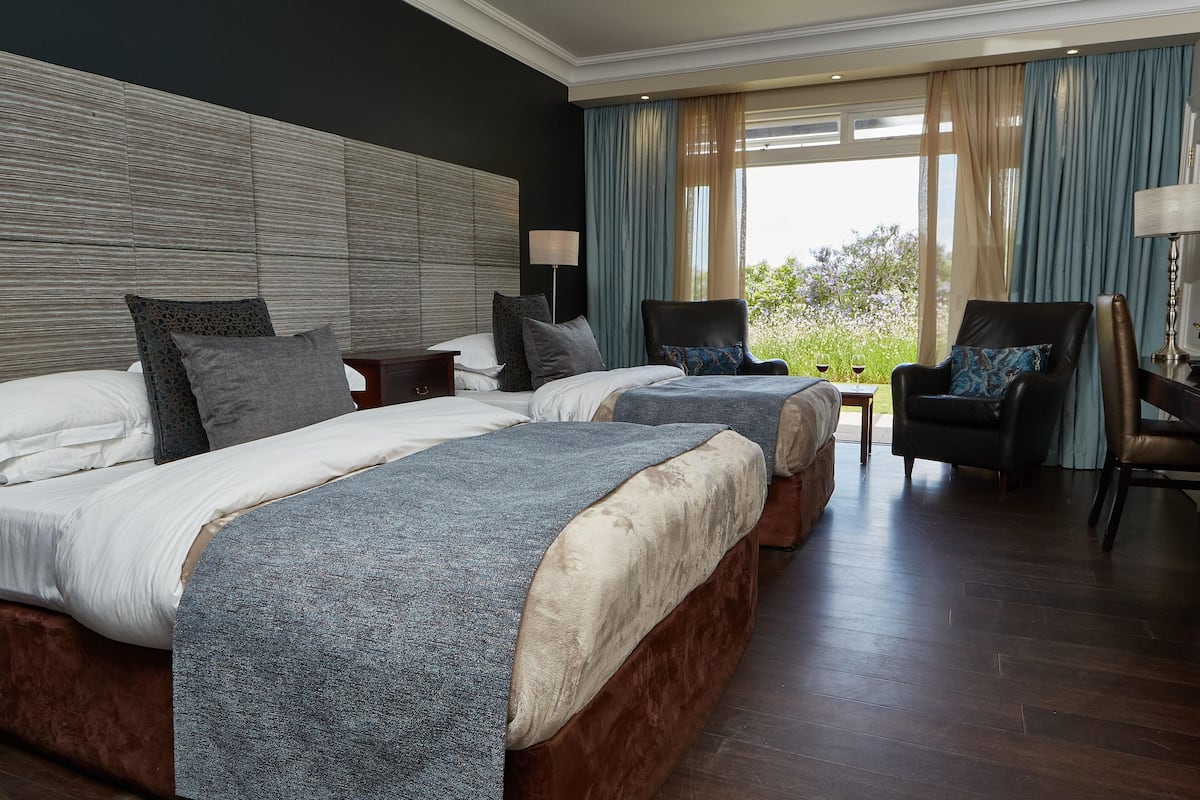 A hotel room with two beds, a large window with curtains, a chair, and a small table with a lamp.