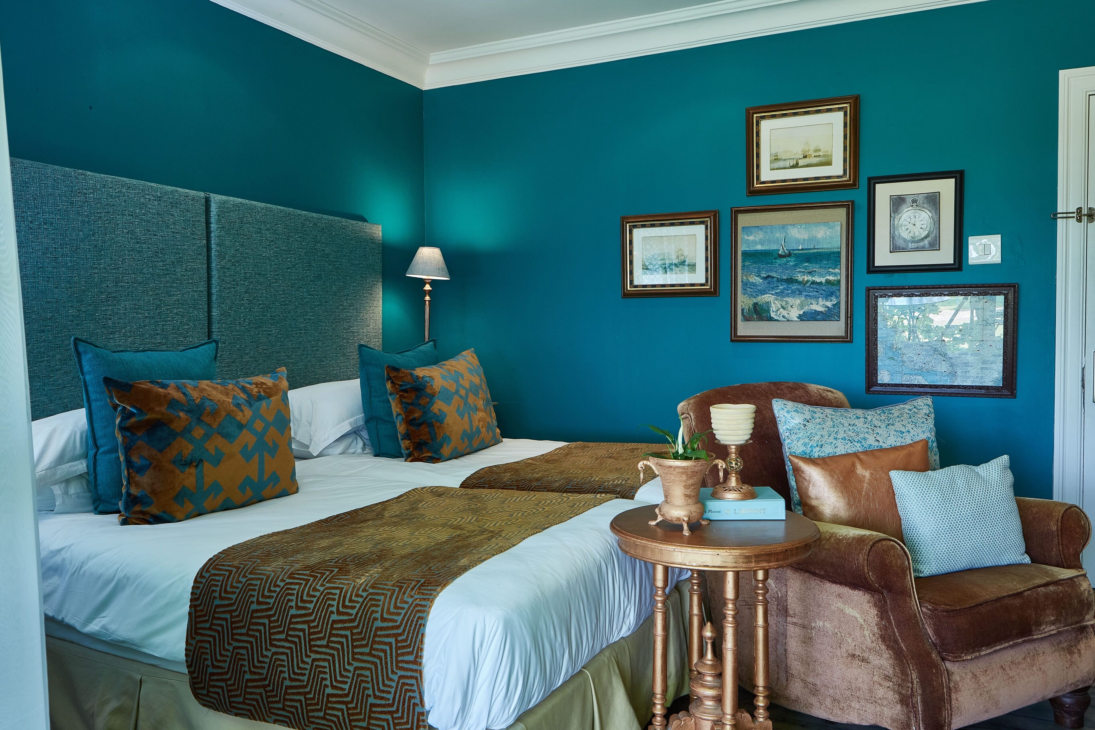 Une chambre d’hôtel avec deux lits, un fauteuil en cuir marron, une petite table ronde et un mur décoré de tableaux encadrés.