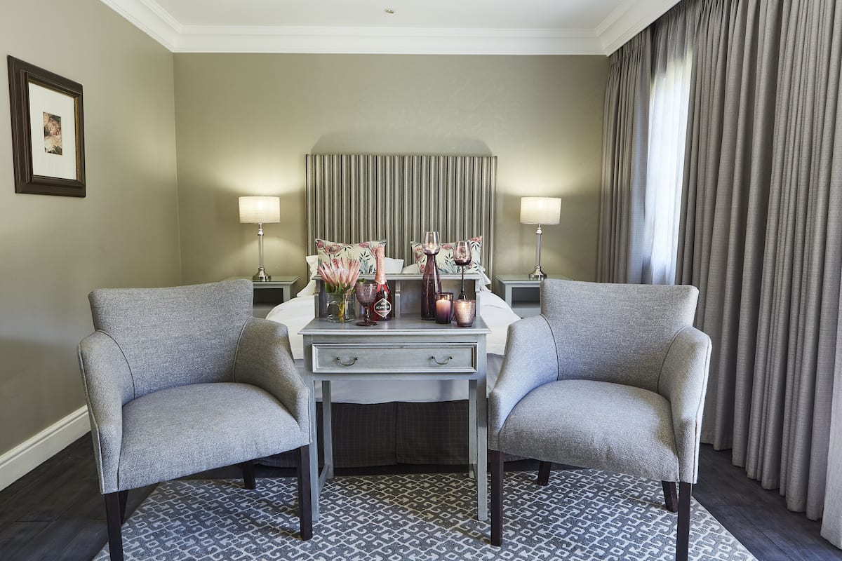 A hotel room with a bed, two armchairs, a small table with decorative items, a lamp, and a window with curtains.