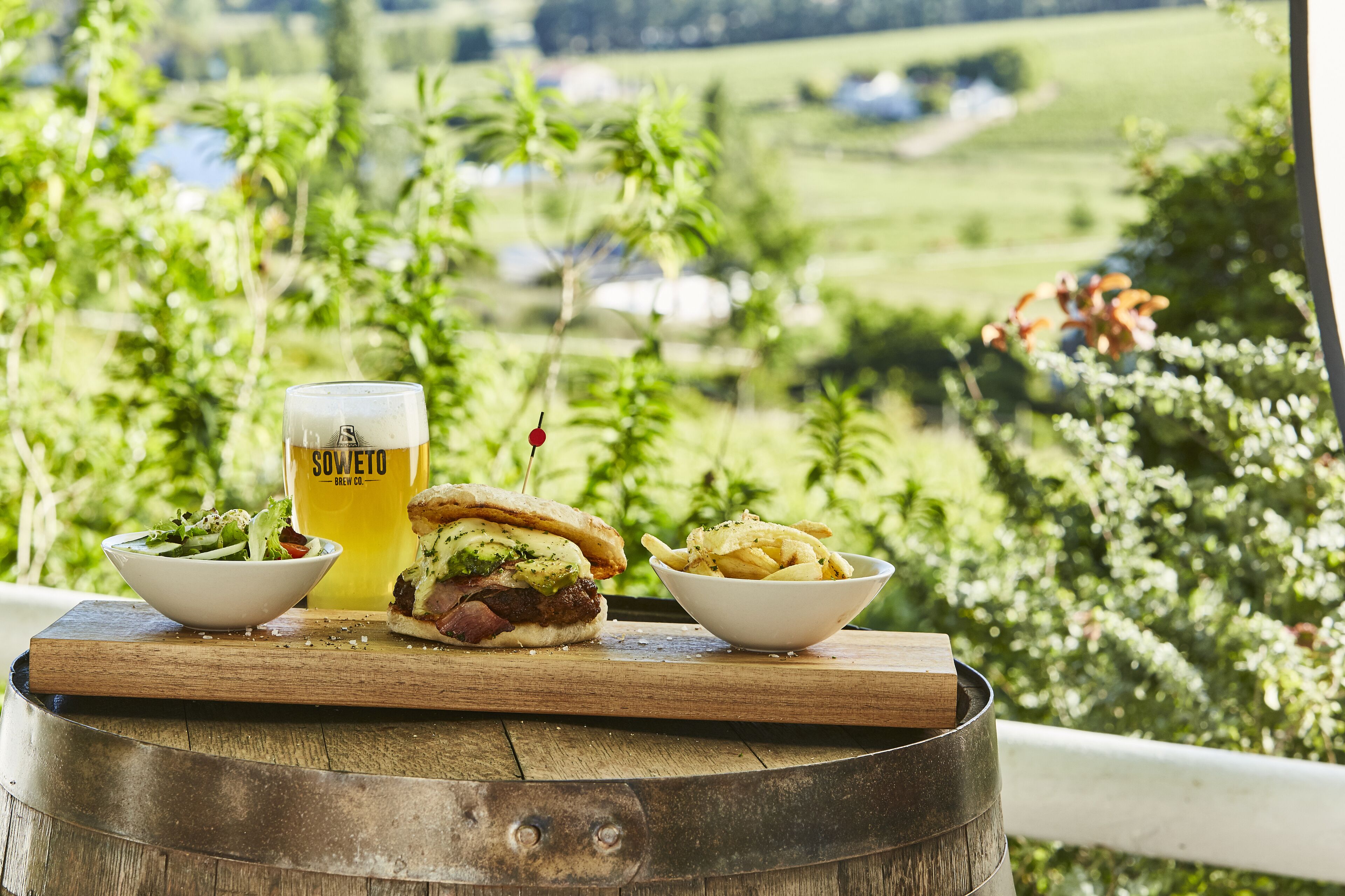 A meal with a sandwich, fries, and a beer on a wooden table with a scenic view.