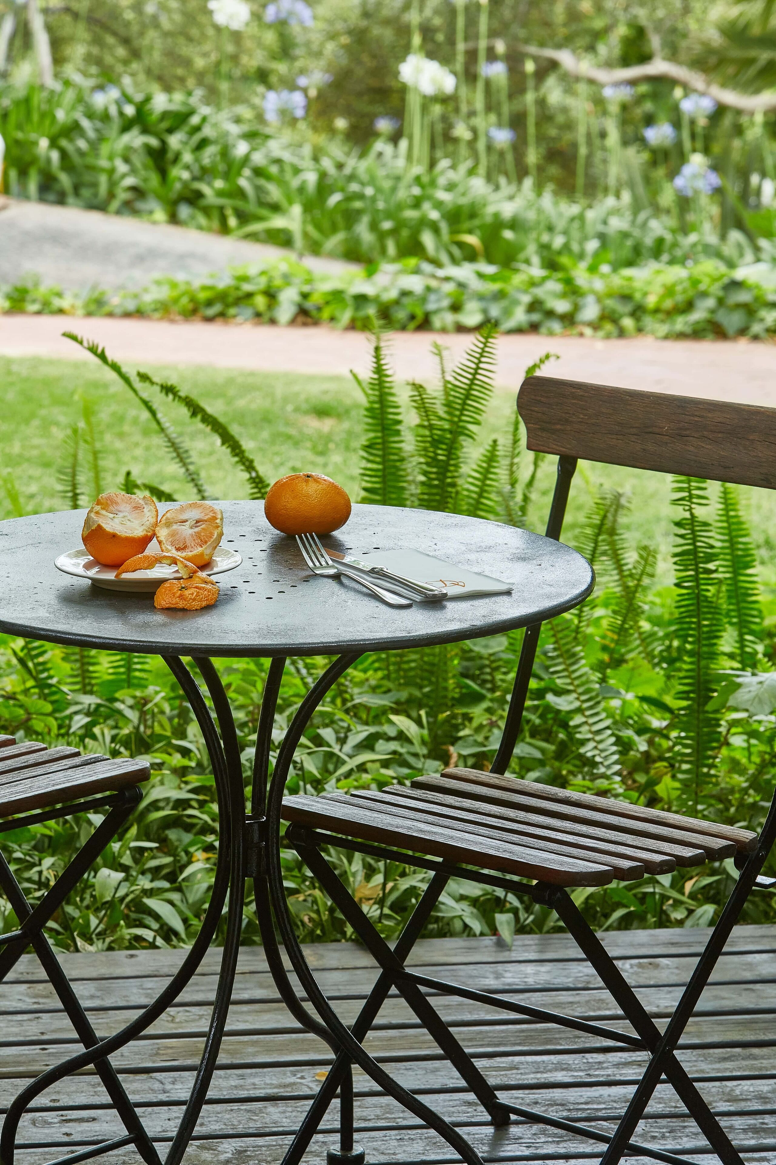 Mesa externa com um prato de laranjas e um caderno, em frente a um jardim exuberante.
