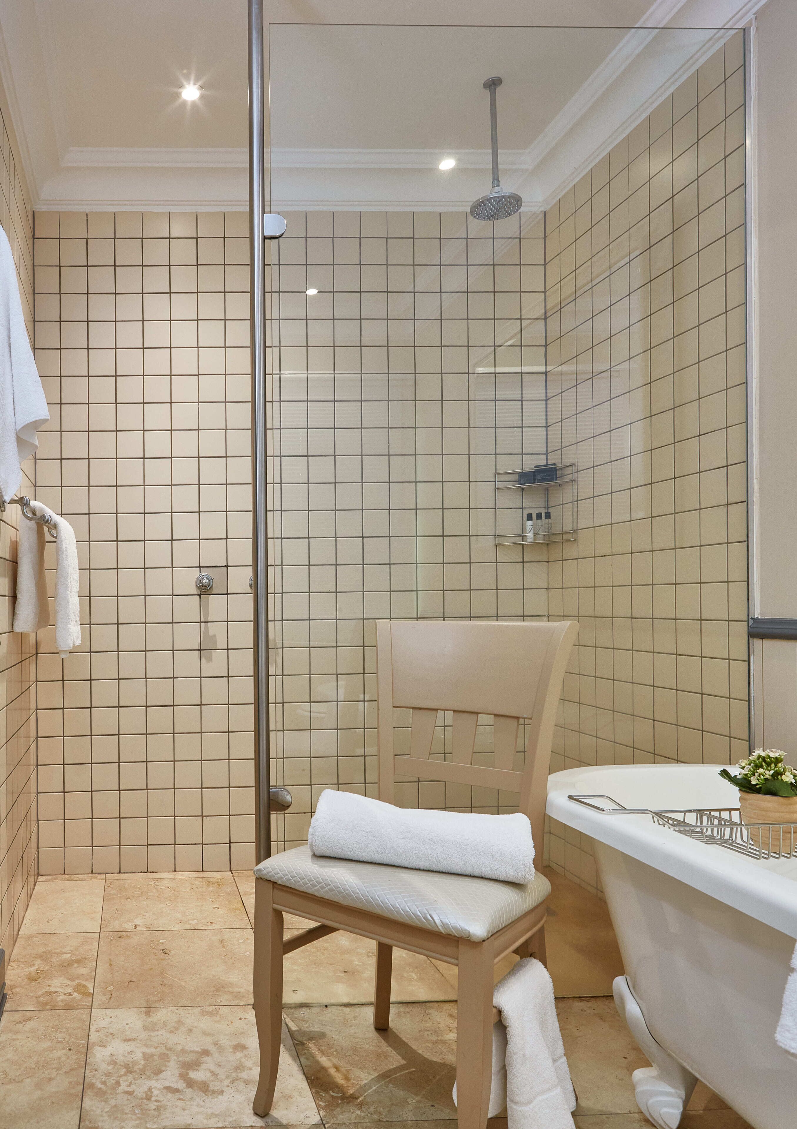 A bathroom with a glass-enclosed shower, a wooden chair, a white bathtub, and a tiled wall.
