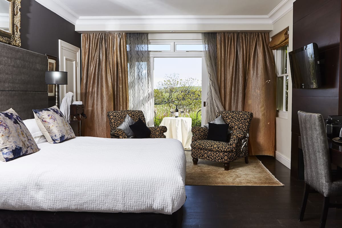 A hotel room with a large bed, two patterned armchairs, a small table, and a TV mounted on the wall.
