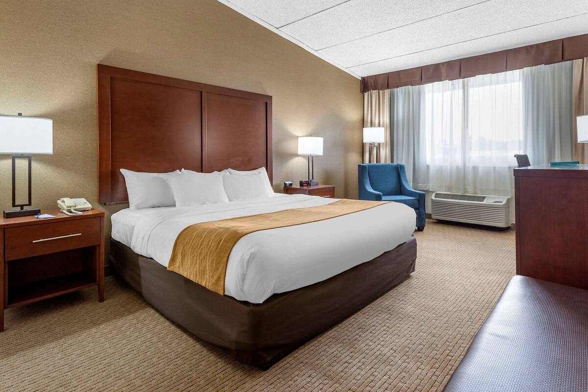 A hotel room with a large bed, two bedside tables with lamps, a chair, and an air conditioning unit.