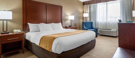 A hotel room with a large bed, two bedside tables with lamps, a chair, and an air conditioning unit.