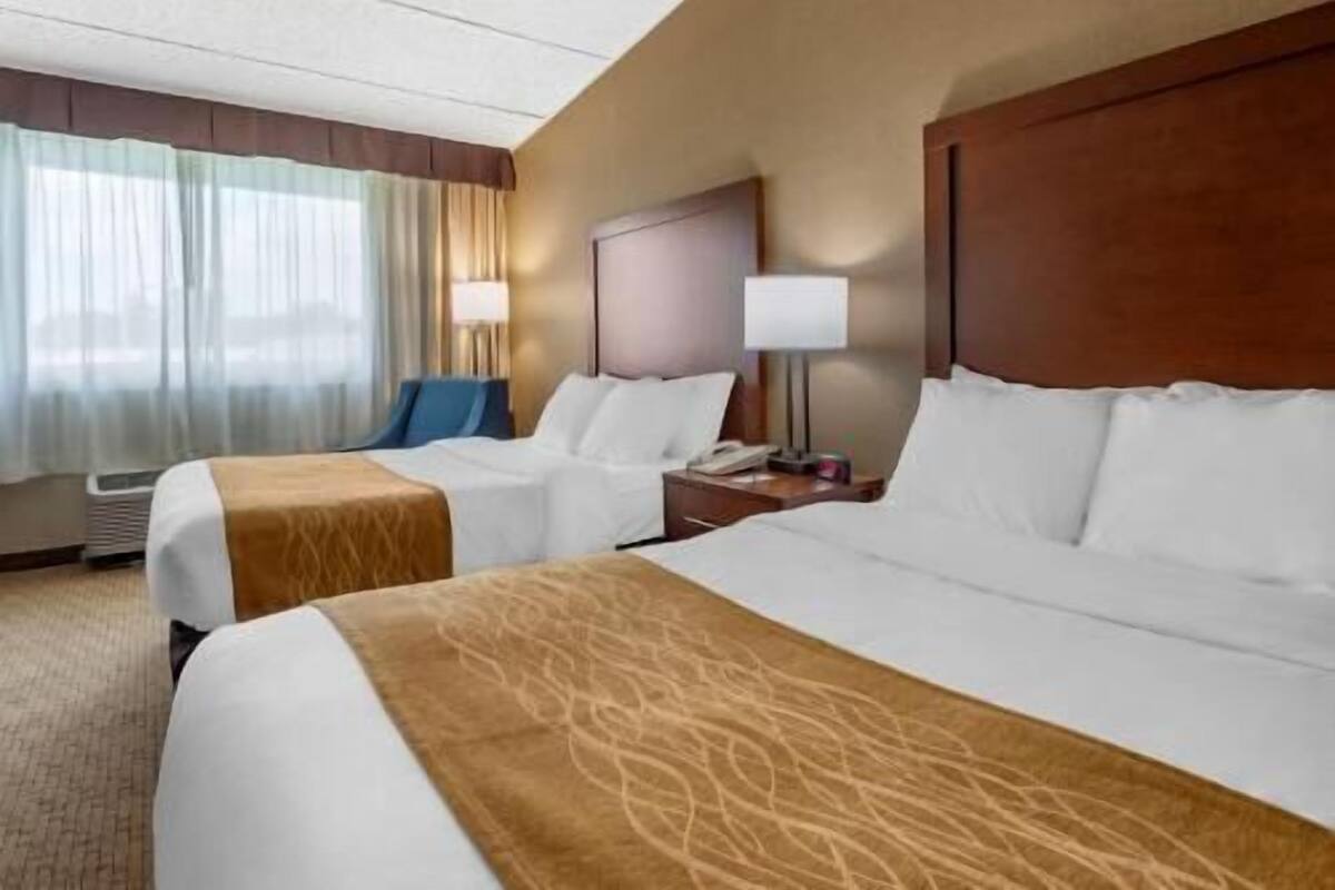 A hotel room with two beds, a nightstand with a lamp, a chair, and a window with curtains.