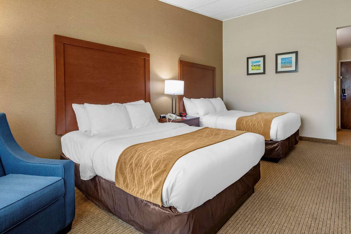 A hotel room with two beds, a blue chair, a lamp, and two framed pictures on the wall.