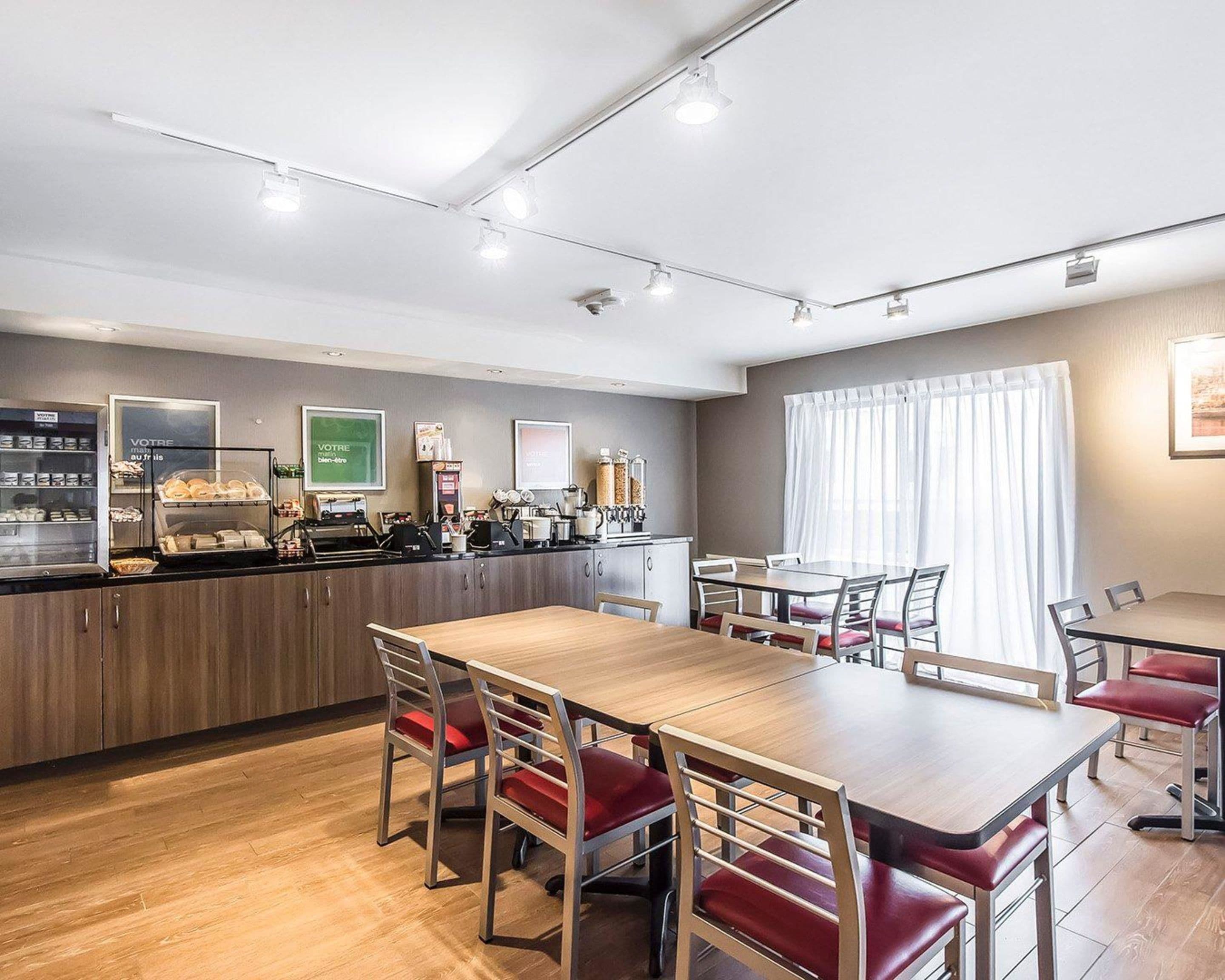 Comedor de hotel con una larga mesa de madera, sillas rojas acolchadas, una barra de café y un amplio ventanal con cortinas.