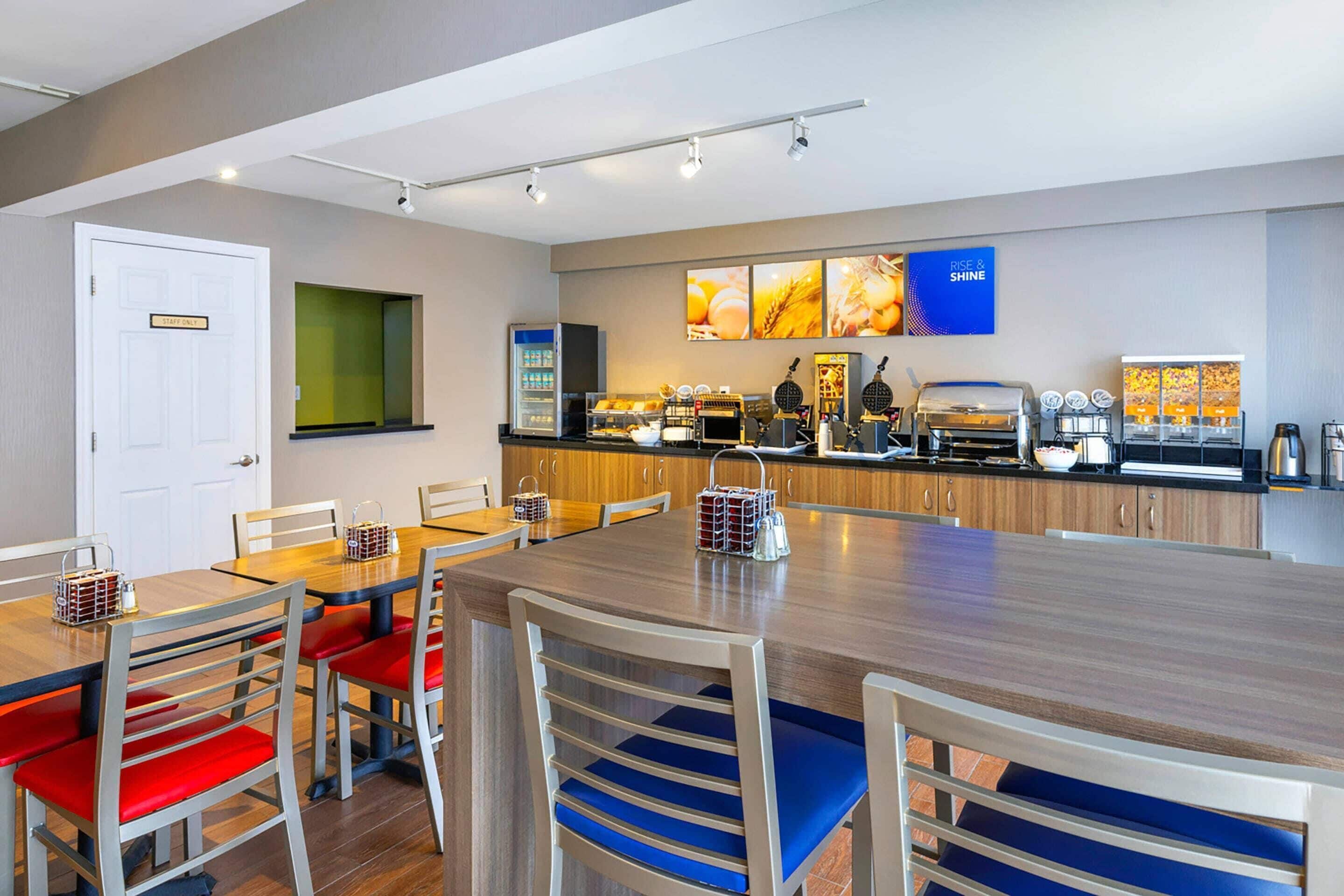 A modern dining area with a wooden table and chairs, a kitchen counter with appliances, and a wall-mounted display.
