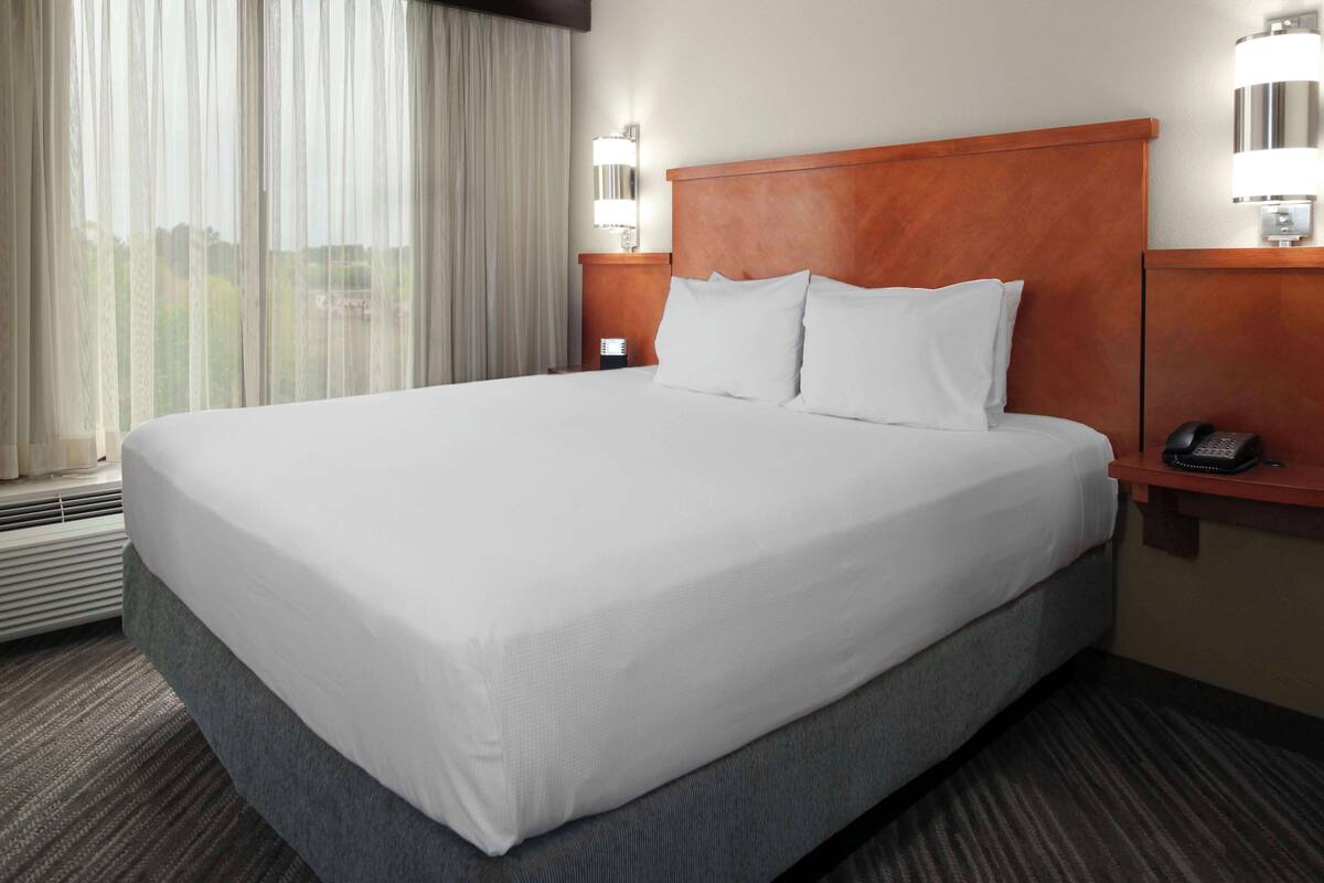 Uma cama grande e bem arrumada com lençóis brancos em um quarto de hotel. Há uma cabeceira de madeira, uma mesa de cabeceira com abajur e um telefone. O quarto tem uma janela grande com cortinas e uma luminária fixada na parede.
