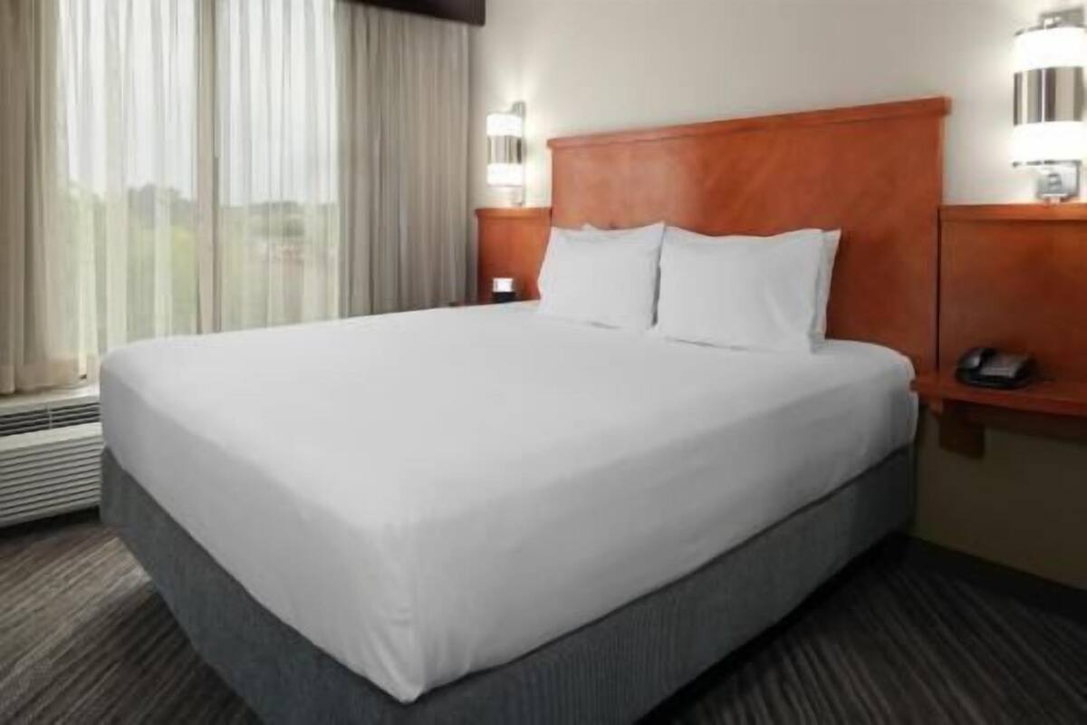 Quarto de hotel com uma cama grande, cabeceira de madeira, mesa de cabeceira com telefone e uma janela com cortinas.