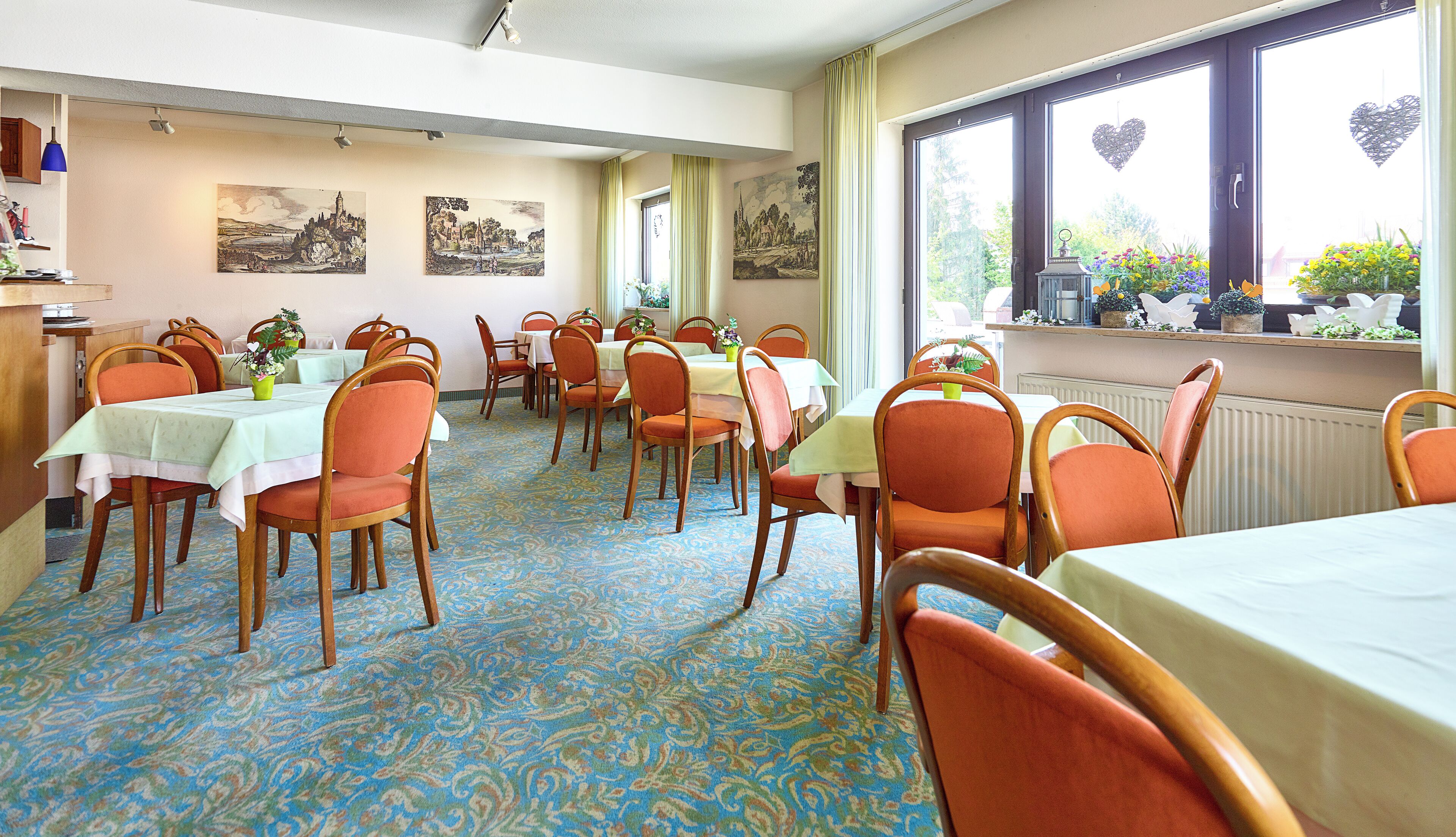 A dining area with tables and chairs, artwork on the walls, and a view of the outdoors through large windows.