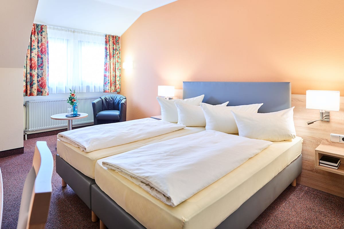 A hotel room with a large bed, bedside tables, a chair, a small table with a vase, and a window with curtains.