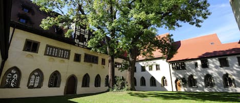 A courtyard with a large tree, a building with arched windows, and a red-roofed structure.