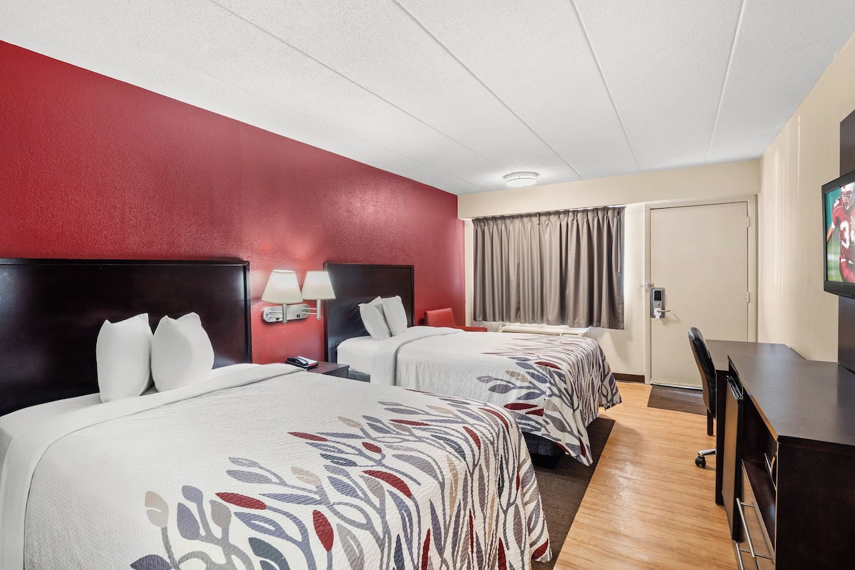 A hotel room with two beds, a desk, a TV, a lamp, and a window with curtains.