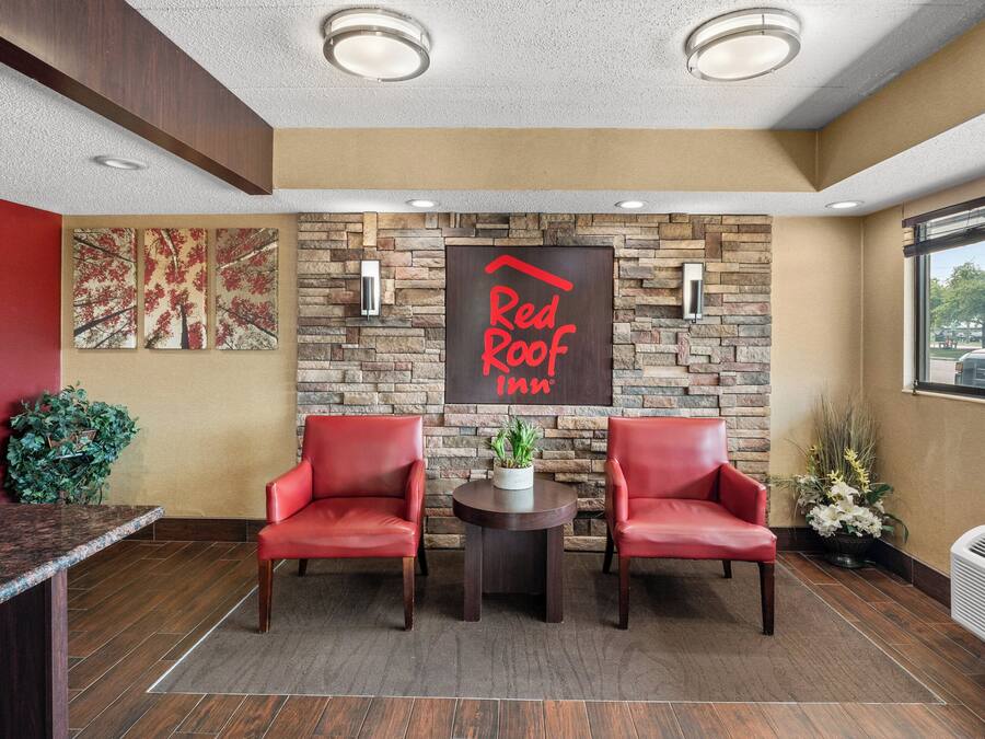 Een receptiegedeelte met een stenen muur, een Red Roof Inn bord, twee rode stoelen, een kleine tafel met een plant en een raam met uitzicht.