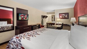 A hotel room with a large bed, a desk with a chair, a television, a desk lamp, and a small table with a lamp. - Red Roof Inn Lansing East – MSU (Lansing)
