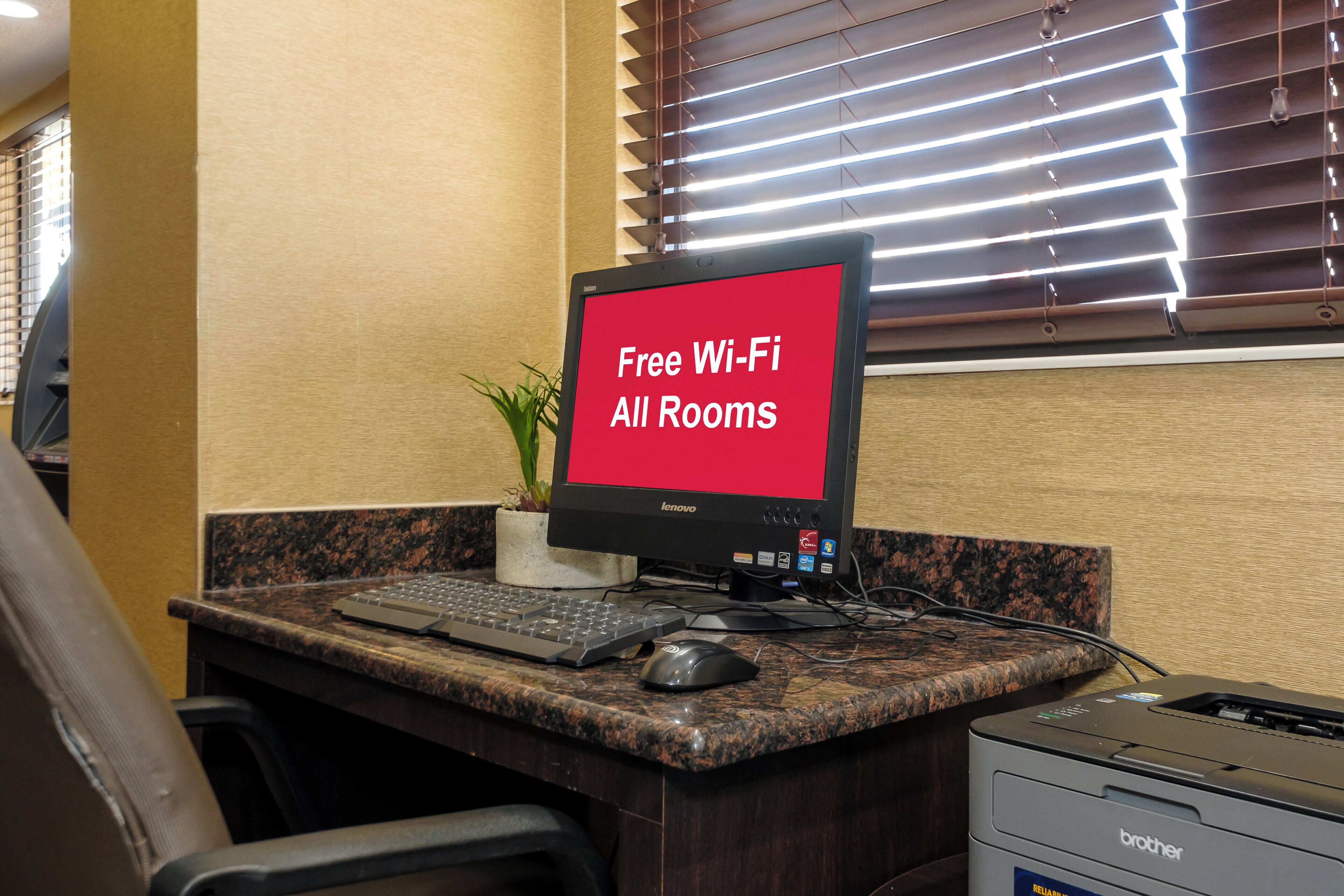 Mesa com monitor de computador exibindo 'Wi-Fi Gratuito em Todos os Quartos', teclado, mouse, impressora e planta em vaso.