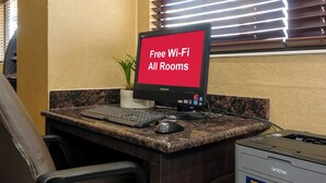 A desk with a computer monitor displaying 'Free Wi-Fi All Rooms,' a keyboard, a mouse, a printer, and a potted plant. - Red Roof Inn Lansing East – MSU (Lansing)