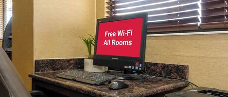A desk with a computer monitor displaying 'Free Wi-Fi All Rooms,' a keyboard, a mouse, a printer, and a potted plant.
