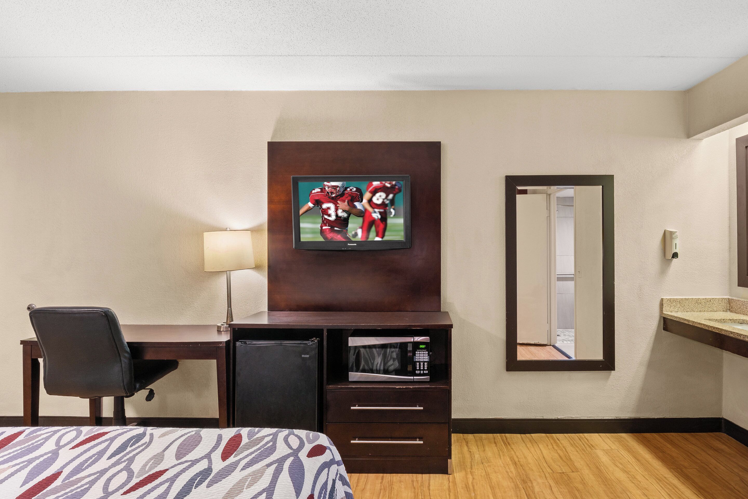 A hotel room with a bed, a desk with a chair, a television mounted on the wall, a microwave, a refrigerator, a mirror, and a sink.