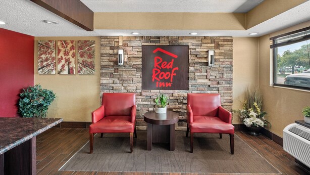 A reception area with a stone wall, a Red Roof Inn sign, two red chairs, a small table with a plant, and a window with a view. - Red Roof Inn Lansing East – MSU (Lansing)