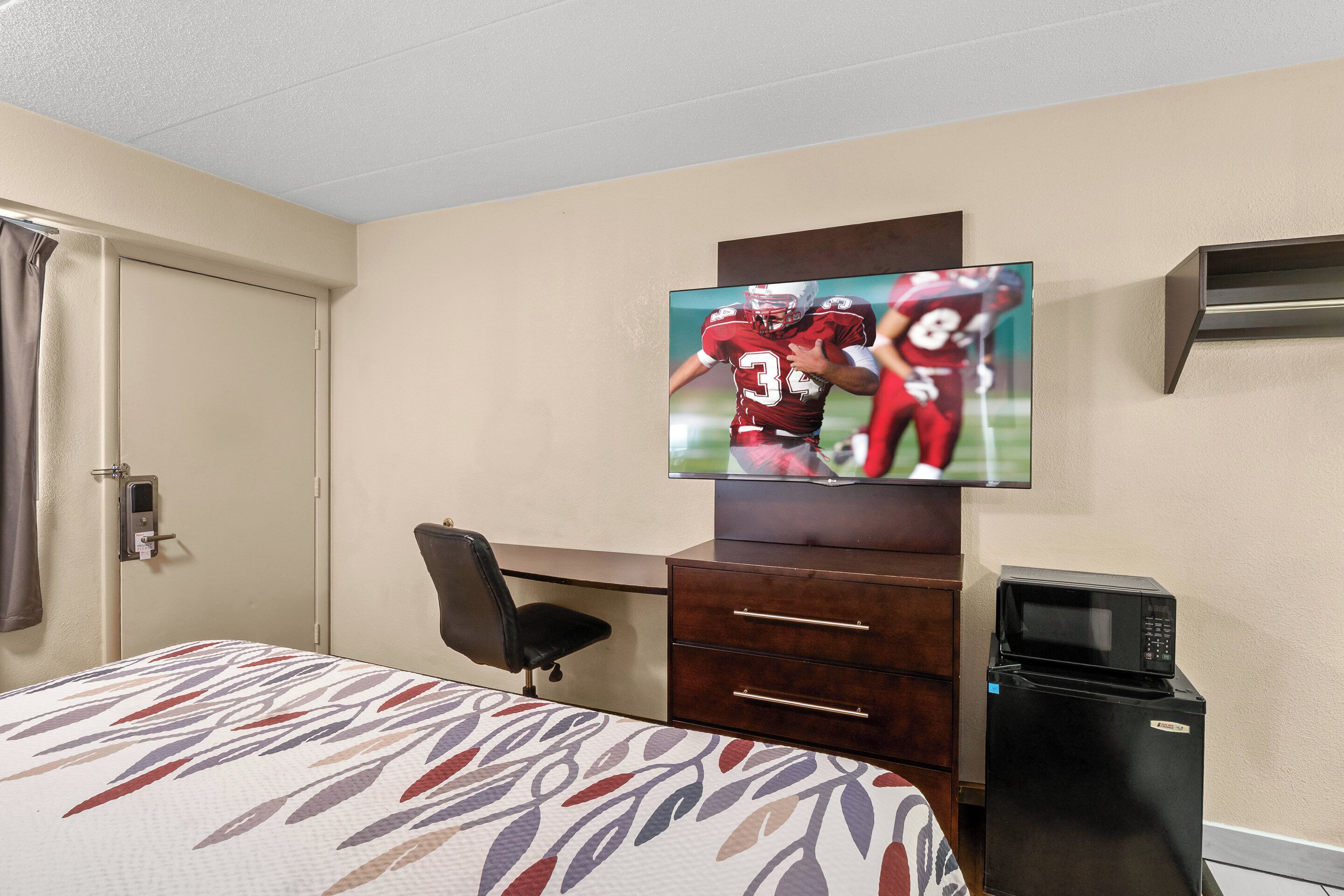 A hotel room with a bed, a desk, a chair, a television mounted on the wall, and a refrigerator.