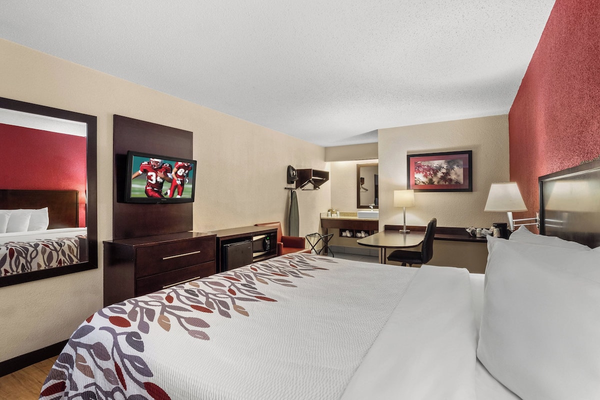 A hotel room with a large bed, a desk with a chair, a television, a desk lamp, and a small table with a lamp.