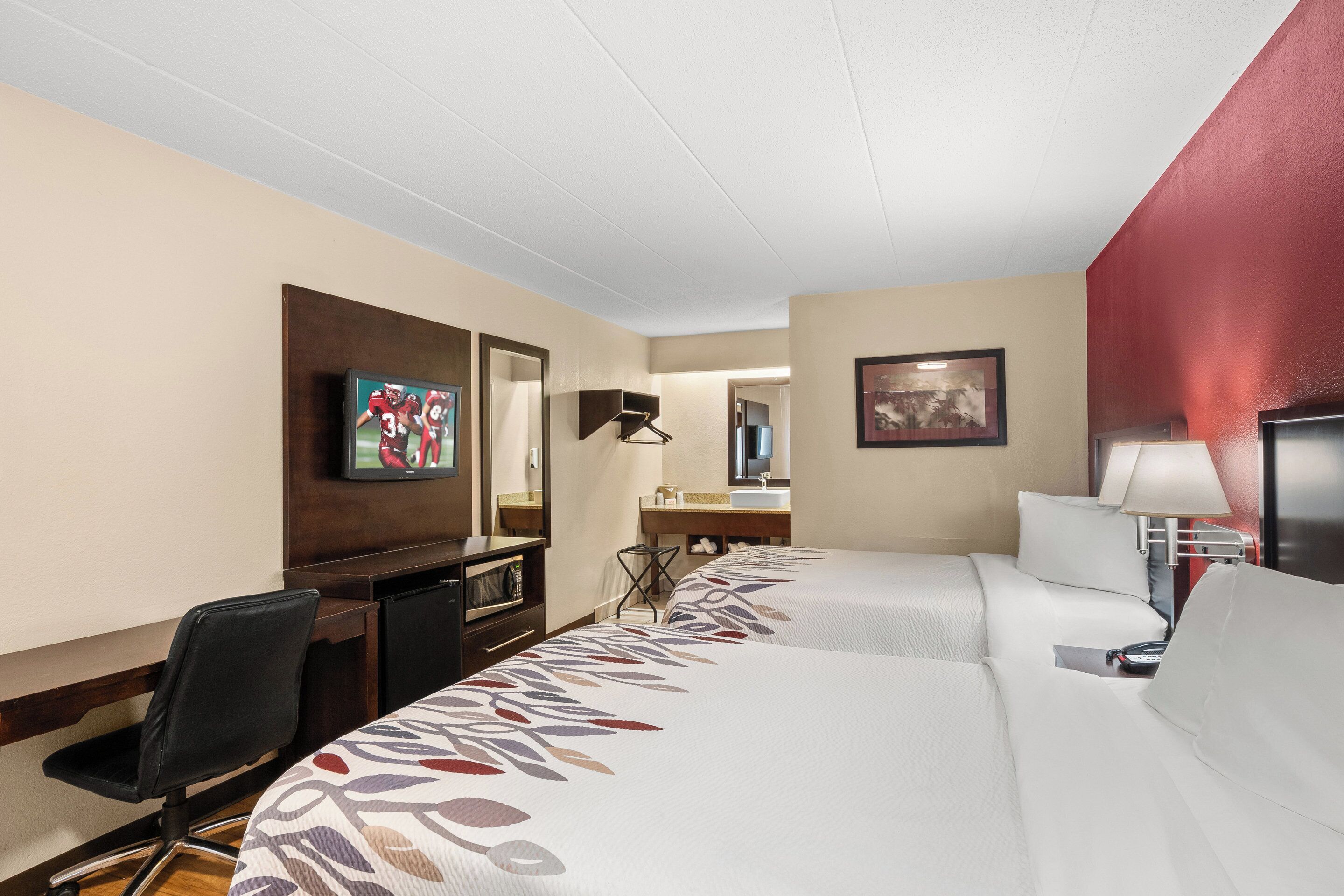 A hotel room with a bed, a desk, a chair, a TV, a mirror, and a bathroom.