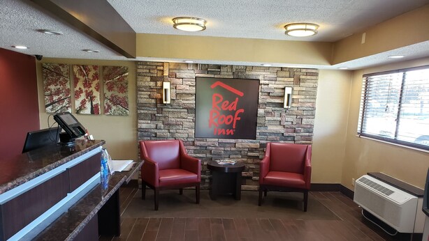 A Red Roof Inn reception area with a stone wall backdrop, two red leather chairs, a reception desk with a payment terminal, and a window with blinds. - Red Roof Inn Lansing East – MSU (Lansing)