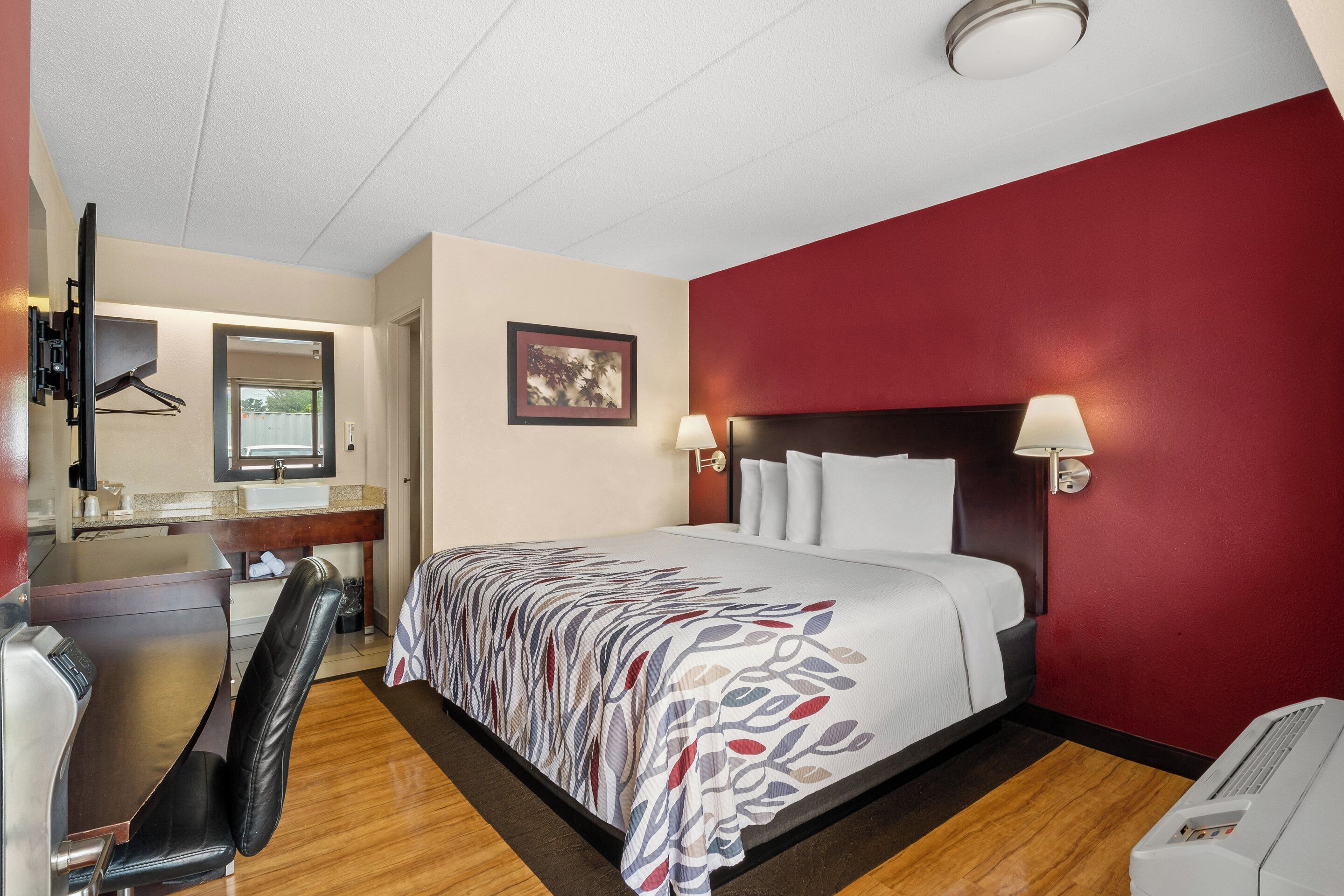A hotel room with a bed, bedside lamps, a desk with a chair, a mirror, and a wall-mounted TV.