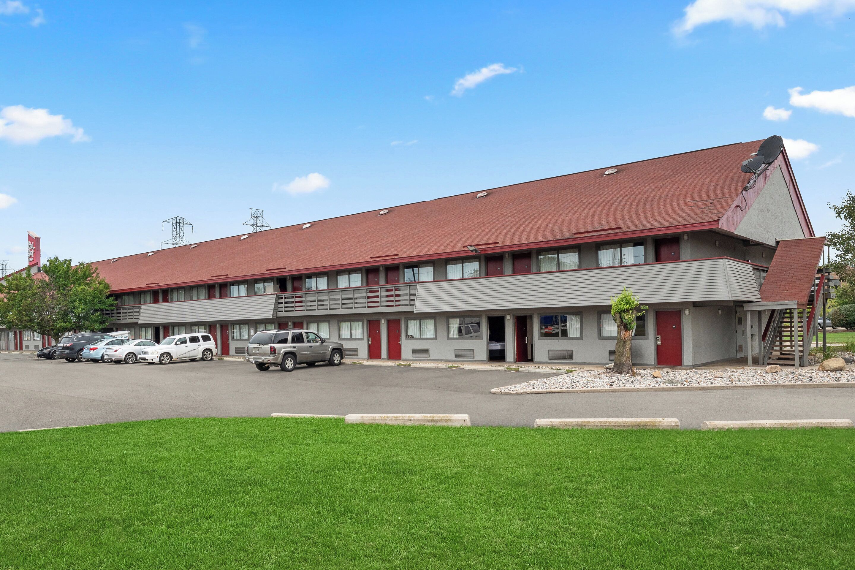 A motel with a red roof, multiple rooms, a parking lot, and a small garden area.