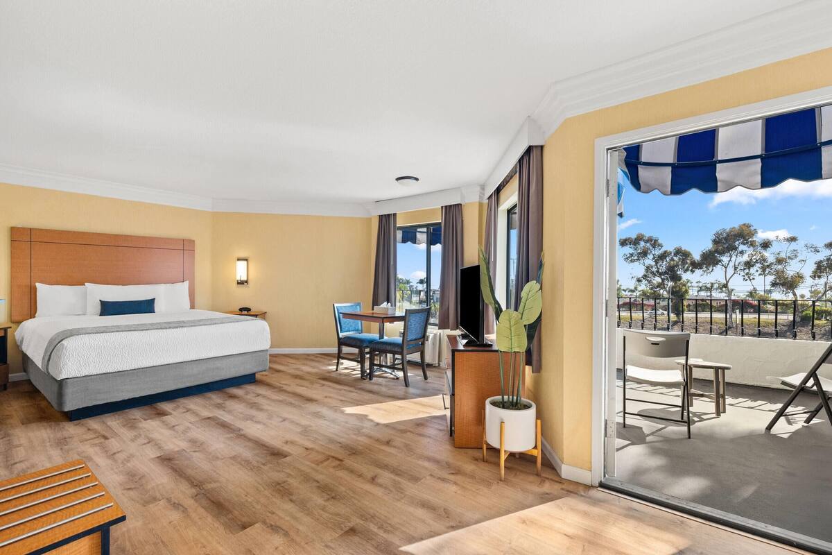 A hotel room with a bed, a desk, a chair, a potted plant, and a balcony with a view.