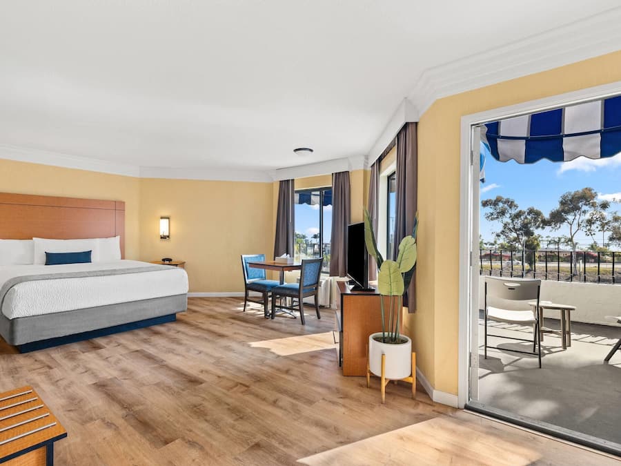 A hotel room with a bed, a desk, a chair, a potted plant, and a balcony with a view.