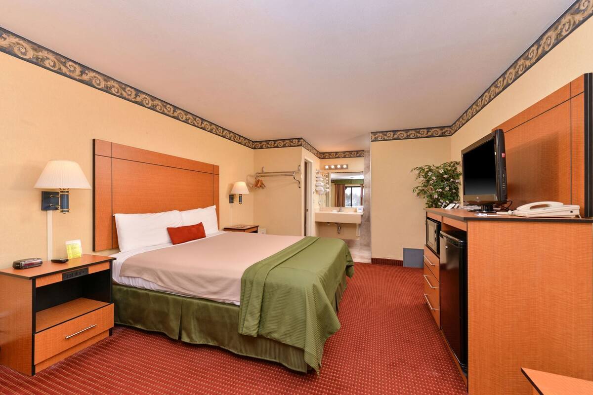 A hotel room with a bed, a desk with a lamp, a television, a desk lamp, a mirror, and a bathroom visible through an open door.