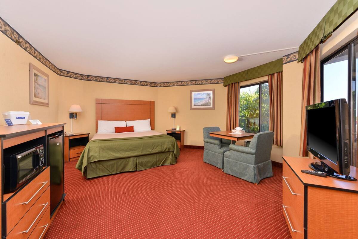 A hotel room with a bed, a desk with a TV, a chair, a small table, a lamp, a nightstand, and a window with curtains.