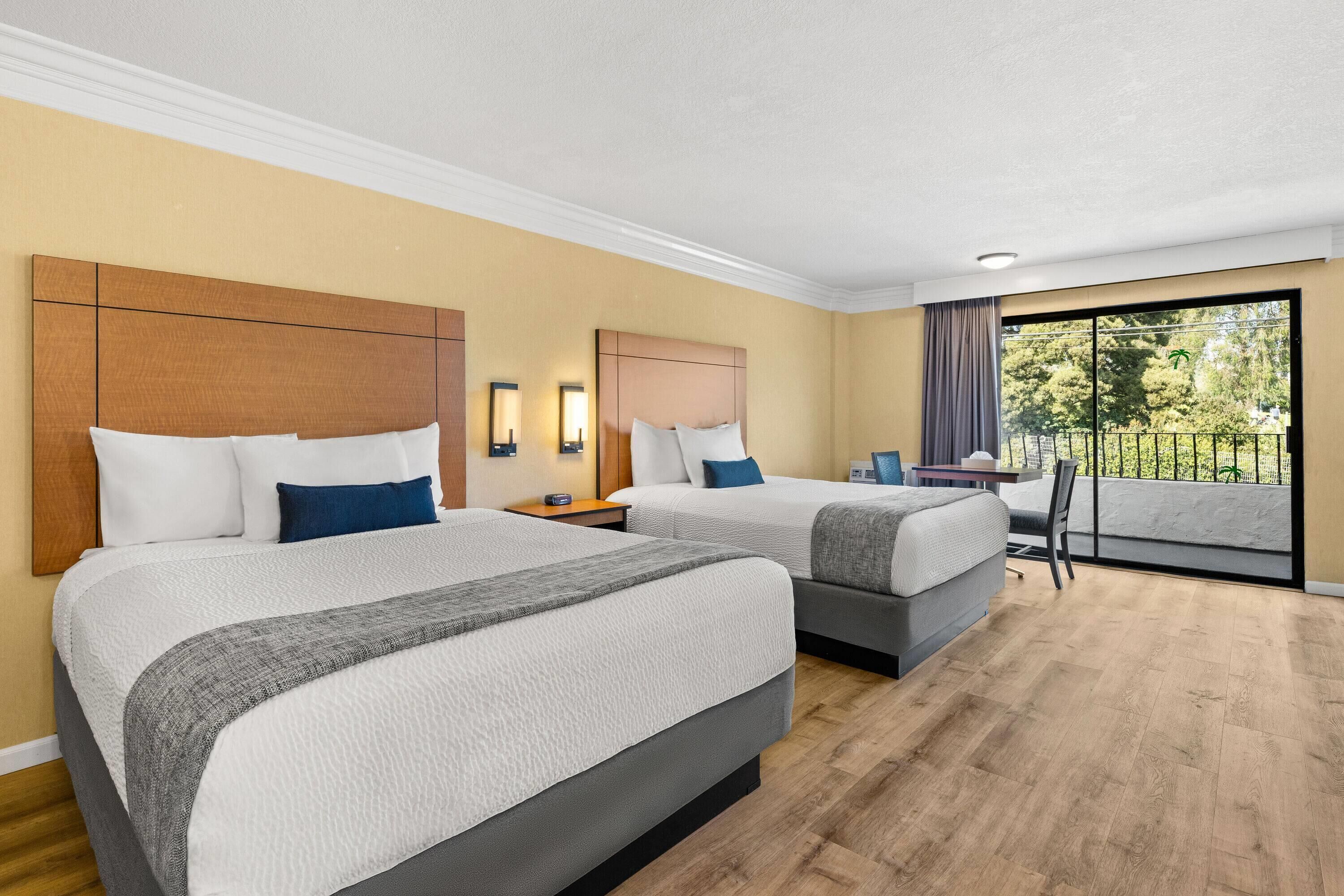 A hotel room with two beds, a desk, and a balcony with a view.