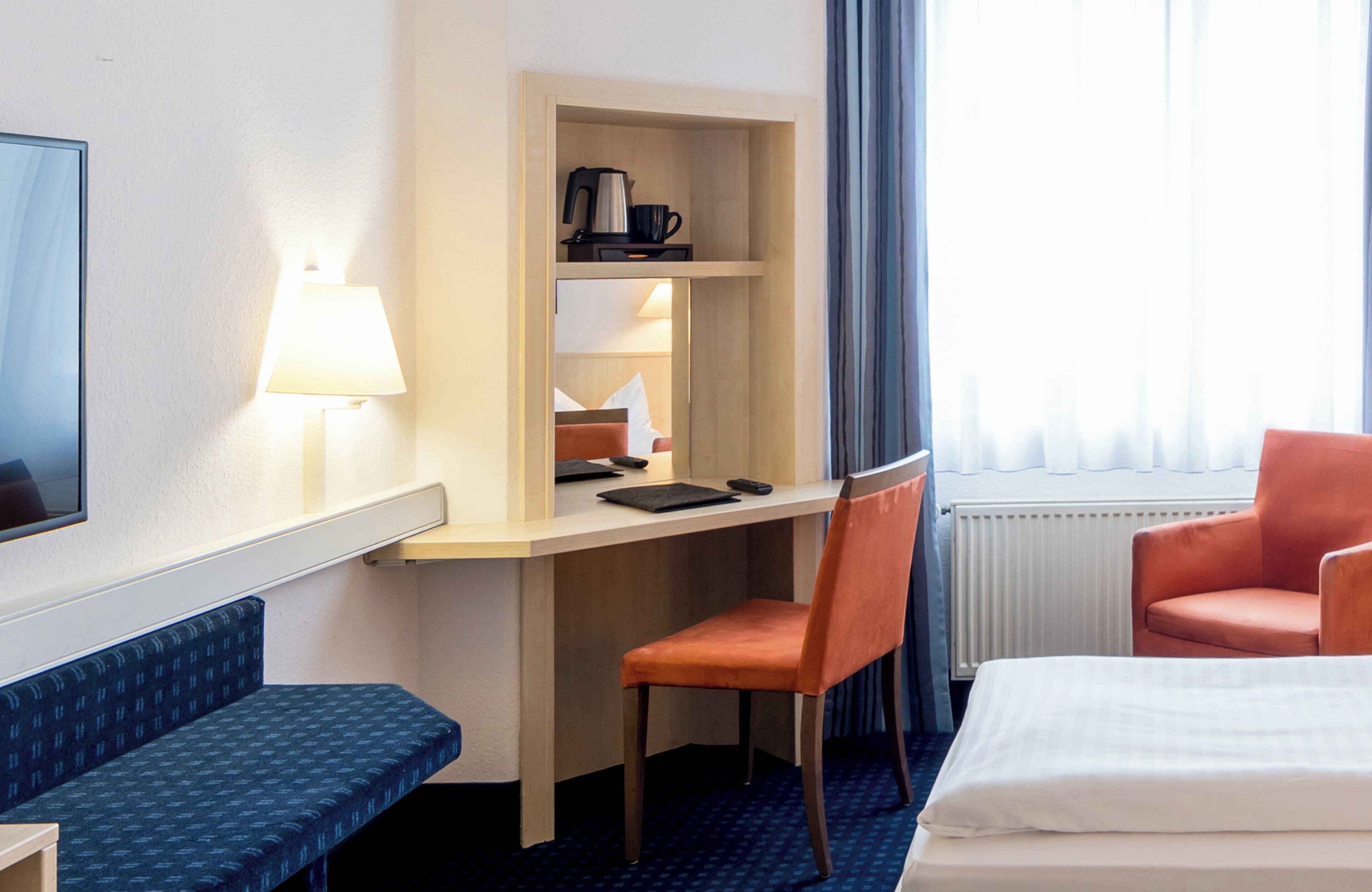 A hotel room with a bed, a desk with a chair, a lamp, a coffee maker, and a window with curtains.