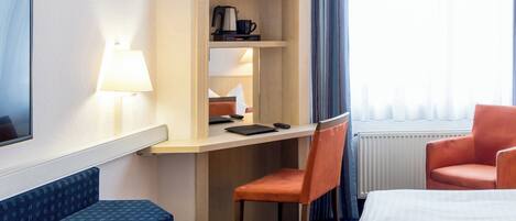 Habitación de hotel con cama, escritorio, silla, lámpara, cafetera y ventana con cortinas.