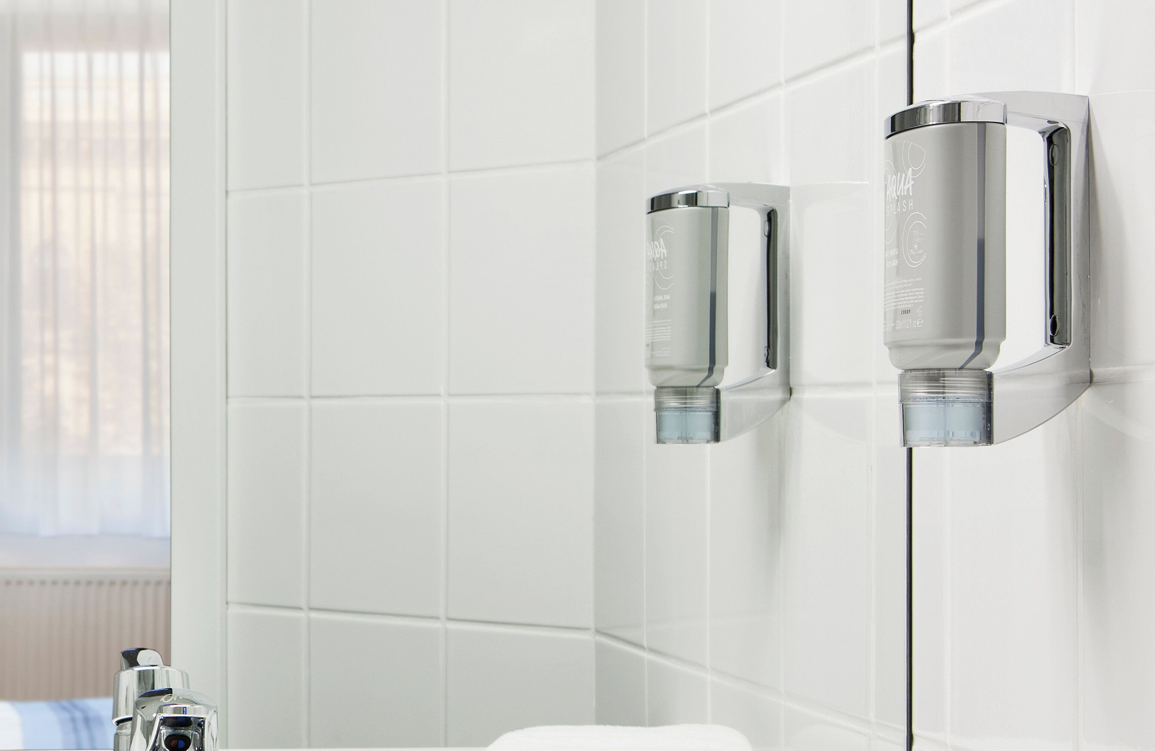 A modern bathroom with white tiled walls, a visible window with blinds, and two chrome fixtures mounted on the wall.