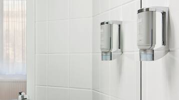 A modern bathroom with white tiled walls, a visible window with blinds, and two chrome fixtures mounted on the wall.